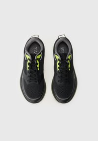Ténis desportivos pretos com detalhes em verde, parte superior em malha, sola texturizada, design com atacadores e colarinho acolchoado. Apresenta orifícios de ventilação para respirabilidade.