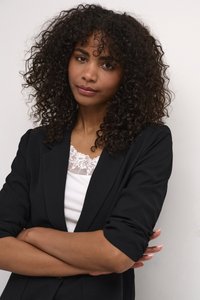 Žena s kudrnatými vlasy, oblečená v černém saku přes bílý krajkový top, stojící se založenýma rukama před světlou stěnou.
