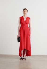 Robe sans manches rouge avec un décolleté en V et une ceinture à la taille, présentant un ourlet asymétrique. Portée avec des sandales noires et tenant un petit sac noir.