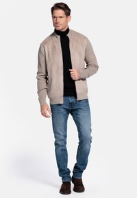 Suède bomberjack in lichtbeige met een gebreide kraag, gecombineerd met blauwe spijkerbroek en bruine suède laarzen. Voorzijde met ritssluiting, textuurrijke afwerking.