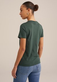 Zeleno kratkotrajno majico iz bombažne tkanine, prilegajočega kroja z okroglo ovratnico. Zadnji pogled razkriva gladko teksturo in subtilne šive.