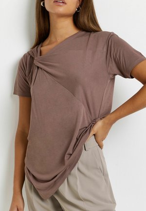 Brown short-sleeve T-shirt with a twisted front design, soft texture, and a draped hem, worn with light beige pants.