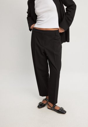 Persona que lleva pantalones negros a medida, blazer negro, top blanco sin mangas y zapatos planos negros con detalles de hebilla y correa.