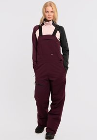 Bordeauxkleurige geïsoleerde overall met verstelbare zwarte banden, een voorzak, een zijritszak en versterkte knieën, gedragen over een roze en zwarte top.