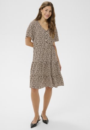 Kobieta uśmiechająca się, ubrana w sukienkę o długości do kolan w panterkę, z krótkimi rękawami i czarne, spiczaste buty na obcasie typu slingback, stojąca na jednolitym tle.