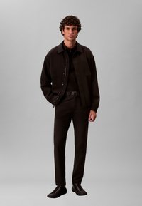 Chaqueta negra con botones a presión, llevada sobre una camisa negra, combinada con pantalones negros y zapatos negros. El modelo está de pie frente a un fondo gris.