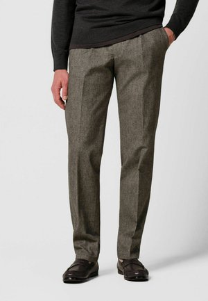 Man draagt grijze pantalon met steekzakken, zwarte loafers en een donker overhemd met lange mouwen, staat met één hand in de zak.