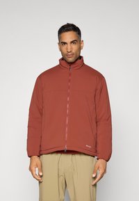 Doudoune bordeaux avec col montant, poches latérales et fermeture éclair. Texture lisse, coupe légèrement ample et petit détail de marque près de l'ourlet.