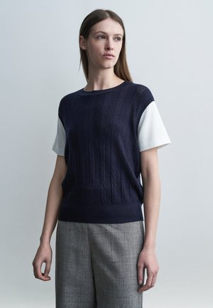 Femeie cu păr brun lung, purtând un pulover cu mâneci scurte bleumarin peste o cămașă albă și pantaloni gri, stând în picioare pe un fundal simplu.