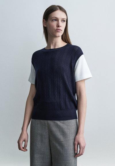 Femme aux longs cheveux bruns portant un pull à manches courtes bleu marine par-dessus une chemise blanche et un pantalon gris, debout devant un fond uni.