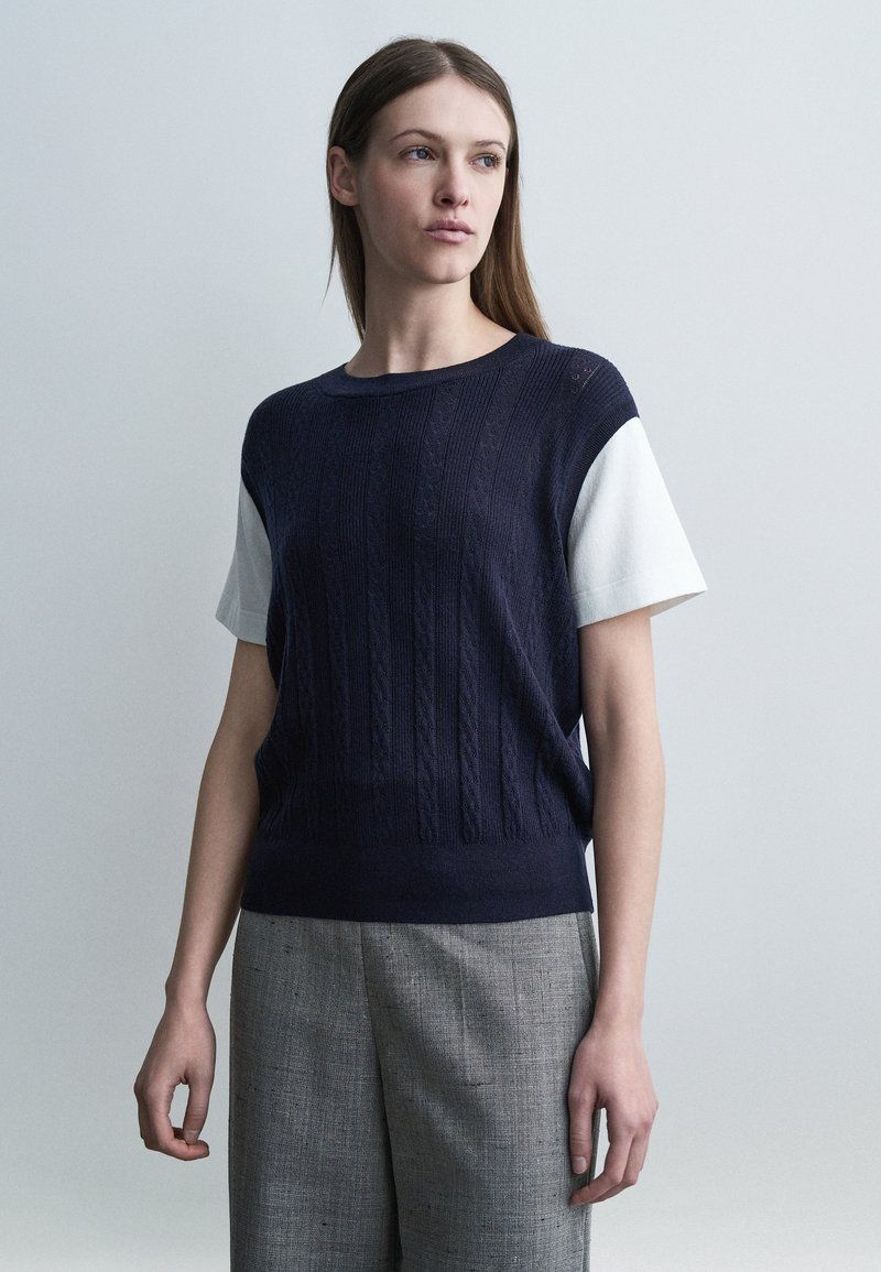 Γυναίκα με μακριά καστανά μαλλιά, φορώντας ένα ναυτικό μπλε πουλόβερ με κοντά μανίκια πάνω από ένα λευκό πουκάμισο και γκρι παντελόνι, στέκεται μπροστά σε απλό φόντο.