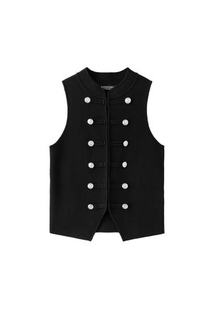 Jeune femme debout, les mains dans le dos, portant un gilet noir sans manches boutonné, un short noir, des chaussettes blanches et des mocassins noirs sur fond gris.
