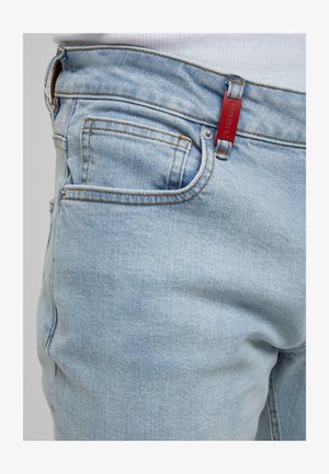 Light blue denim jeans with orange stitching and a small red rectangular tag attached to the waistband near the front pocket.