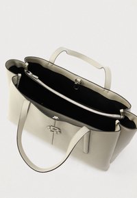 Beżowa skórzana torba typu tote z ciemnym wnętrzem, wyposażona w dwa solidne uchwyty oraz zapinaną na zamek kieszeń. Zawiera ozdobny element z metalu.