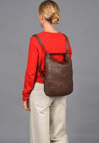 Bruine geweven leren rugzak met verstelbare riemen, met een slouchy vorm en een gestructureerd oppervlak. Model draagt een rode trui en lichte broeken.