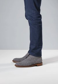 Grijze suède chukka laarzen met een ronde neus, lichtbruine zool en minimale veters, gecombineerd met donkerblauwe broek in een staande houding.