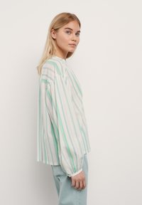 Chemise rayée en vert clair et blanc, avec des manches bouffantes, un col rond et une coupe décontractée. Portée avec un pantalon bleu clair.