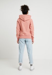 Vaaleanpunainen huppari, jossa on keskellä selkää oleva sauma ja kiristysnauhalla varustettu huppu, yhdistetty vaaleansinisillä, resoreilla varustetuilla farkuilla ja valkoisilla lenkkareilla, joissa on mustia yksityiskohtia.