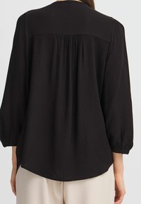 Blouse noire avec des manches ballon et un tissu texturé. Présente un dos plissé et un ourlet arrondi. Le matériau semble léger et fluide.