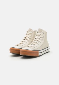 Vysoké tenisky v off-white plátne s gumovou špičkou a hnedou podrážkou. Obsahujú kovové očká a čierny pruh. Textúrovaná tkanina.