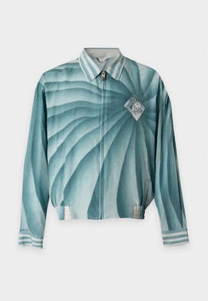 Man met zwarte zonnebril, teal spiraalpatroon ritssluiting jas, zwarte broek en witte sneakers staand tegen een effen achtergrond.