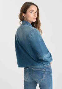 Woman with long brown hair looks over shoulder wearing cropped denim jacket and blue jeans against plain light background.
