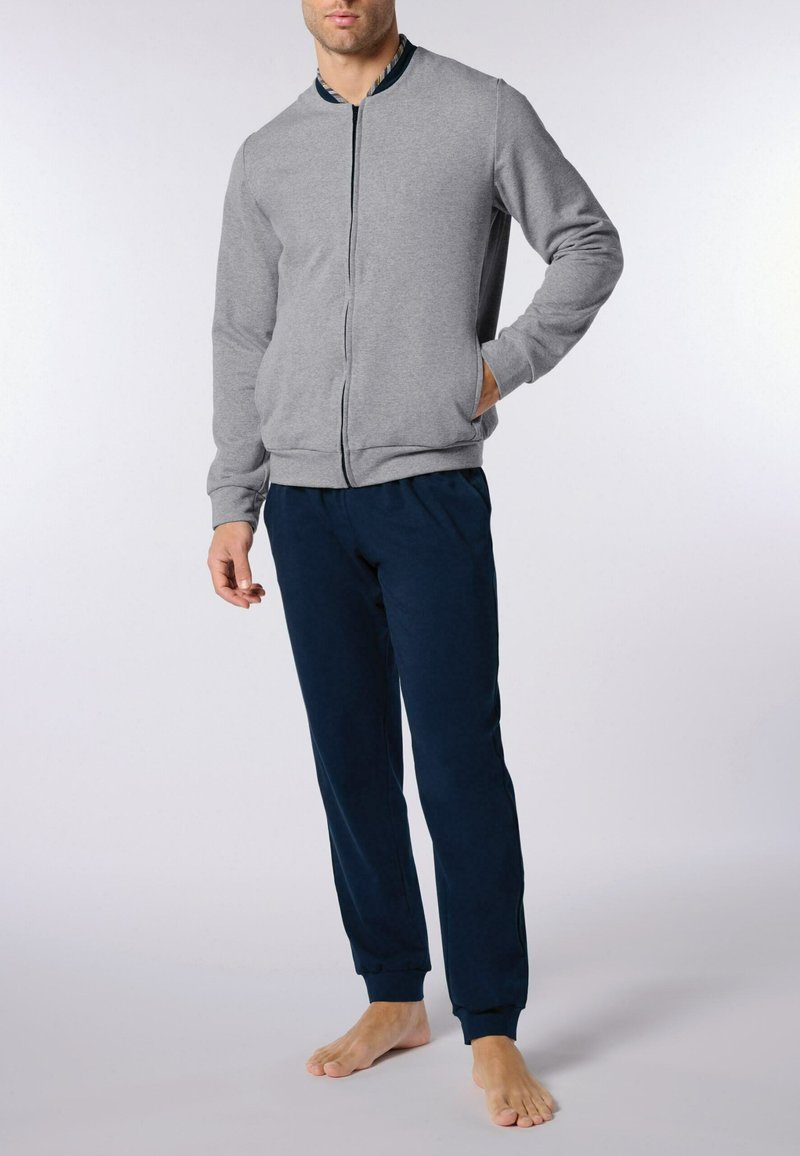 Uomo che indossa una giacca zip grigia e pantaloni jogger blu navy, in piedi a piedi nudi contro uno sfondo chiaro e semplice.