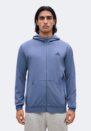 Man met middellang donker haar, die een blauwe zip-up hoodie en lichtgrijze joggingbroek met zwarte logo's draagt, staand tegen een effen achtergrond.