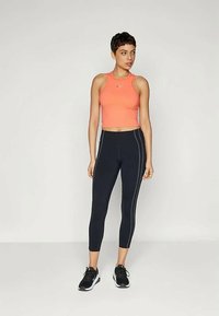 Žena v oranžovom bezrukávovom crop topu a čiernych priliehavých legínach s bielymi švami, stojí pred jednoduchým svetlým pozadím.