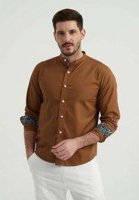 Chemise marron à manches longues avec un col montant, ornée d'accents colorés sur les poignets et de boutons blancs contrastants.