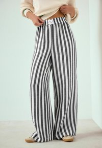 Pantaloni cu croială largă, cu dungi verticale alb-negru, țesătură texturată și talie elastică, asortați cu pantofi slip-on de culoare maro deschis.