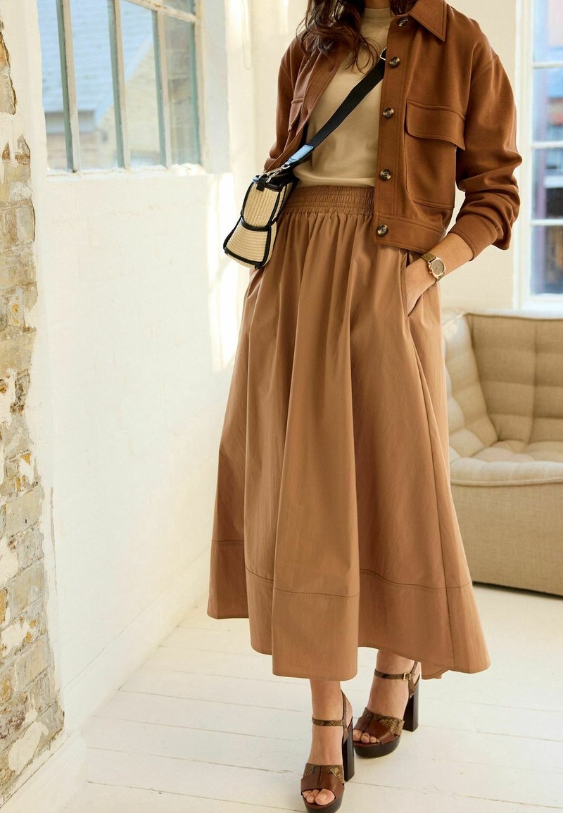 Femme portant une veste marron boutonnée, un haut beige, une longue jupe marron, un sac bandoulière, une montre et des sandales marron à talons hauts, debout à l'intérieur.