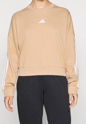 Personne portant un sweat-shirt beige Adidas à manches longues avec des bandes blanches sur les manches et un pantalon noir sur un fond uni.