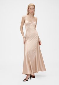 Robe maxi en satin couleur nude clair, avec fines bretelles, corsage ajusté et ourlet évasé. Le mannequin porte des talons noirs à lanières.