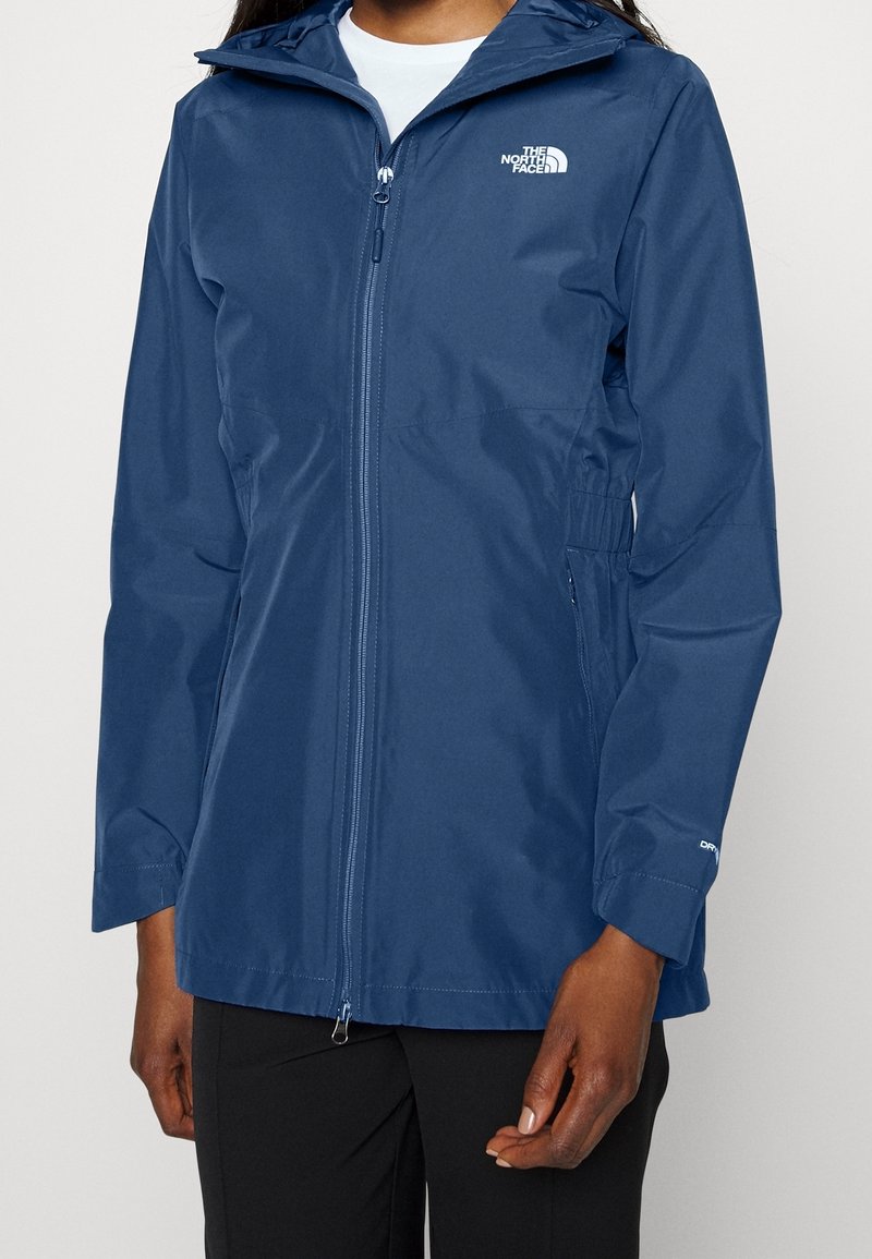 Femme portant une veste imperméable bleu marine avec capuche, fermeture éclair à l'avant et logo The North Face sur la poitrine.