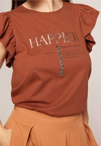 Brun bomull T-shirt med volangärmar, med en metallisk texttryck som säger "HAPPIER" och en paljettdetalj nedanför.