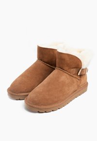 Botas de tobillo de ante marrón con forro de piel de oveja y detalle de correa ajustable. Suela de goma plana, puntera redondeada y textura suave.