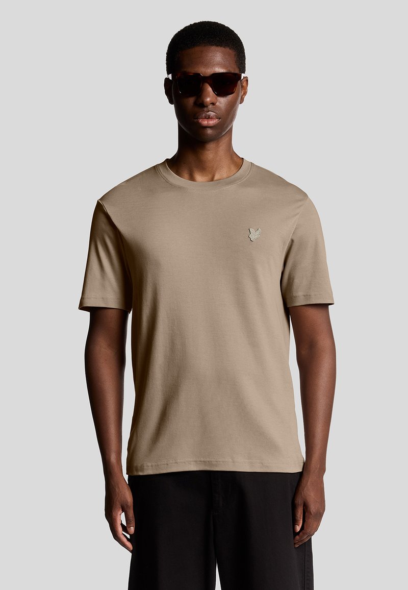 Beige Baumwoll-T-Shirt mit rundem Ausschnitt und kurzen Ärmeln, das ein kleines gesticktes Logo auf der Brust und eine glatte Textur aufweist.