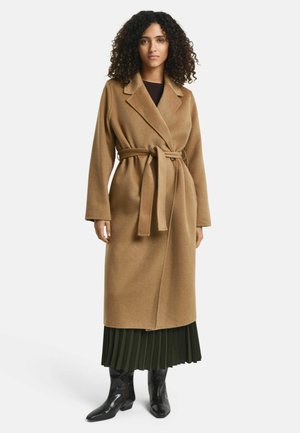 Camel-colored, long wool coat with a belt, notched collar, and long sleeves, layered over a pleated green skirt and black ankle boots.