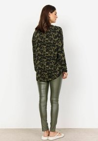 Grön kamouflagemönstrad långärmad topp med rundad nederkant, matchad med åtsittande gröna leggings i konstläder. Enkla sandaler fulländar looken.