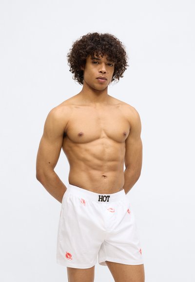 Costume de baie alb cu imprimeuri roșii în formă de sărut, talie elastică cu „HOT” scris în negru, textură netedă și croială lejeră. Modelul stă cu brațele în spate.