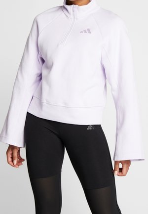 Femme portant un sweat-shirt blanc à fermeture éclair quart de zip avec des manches larges et des leggings noirs Adidas avec des panneaux en maille sur les cuisses.