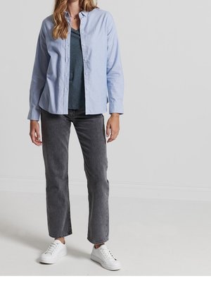 Kobieta w niebieskiej koszuli z guzikami nałożonej na ciemnoszarą koszulkę, szare dżinsy o prostych nogawkach i białe trampki, stoi na tle jednolitego tła.