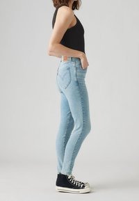 Frau, die hellblaue, hoch sitzende Skinny-Jeans mit herzförmigen Rückentaschen-Nähten und schwarzen High-Top-Sneakers trägt, steht seitlich.