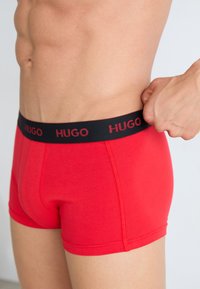 Piros pamut boxer alsónemű fekete derékszíjjal, amelyen a "HUGO" piros betűkkel szerepel. Feszes fazonnal és sima textúrával rendelkezik.