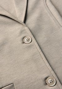Gros plan d’un tissu beige texturé avec deux boutons beiges cousus, montrant les coutures et les bords de tissu qui se chevauchent.
