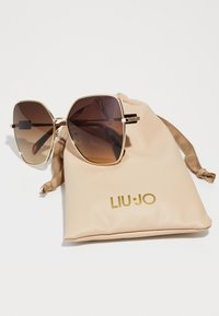 Occhiali da sole con lenti esagonali marroni oversize e montatura in metallo dorato, poggiati su un sacchetto con coulisse beige contrassegnato "LIU·JO" in oro.