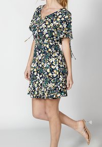 Žena v šatech s krátkým rukávem a květinovým vzorem na tmavém pozadí, s průhlednými sandály na podpatku, pózuje s jednou nohou mírně zvednutou.