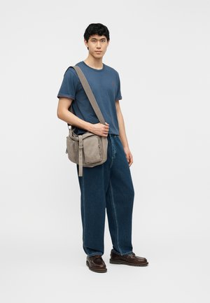 Tânăr purtând un tricou albastru și blugi largi, având o geantă de umăr bej, stând pe un fundal simplu și deschis la culoare.