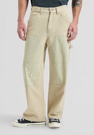 Uomo con pantaloni da lavoro beige a taglio largo con toppe rinforzate sulle ginocchia e sneakers nere basse, in piedi davanti a uno sfondo neutro.